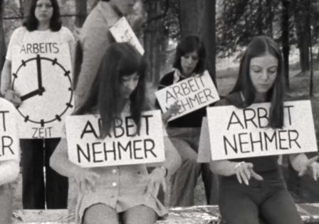 Schwarz-wei&szlig;-Szenenfoto: Junge frauen sitzen auf einer Bank in der Natur, sie machen Tipp-Bewegungen mit den H&auml;nden, als w&uuml;rden sie Schreibmaschine schreiben. Um den Hals haben h&auml;ngen Schilder auf denen das Wort Arbeitnehmer steht.