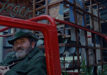 Dein Mann mit gr&uuml;nem Hut und Bart sitzt in einem roten Bus, im Hintergrund ein Haus mit einem Portr&auml;t Erich Honeckers.