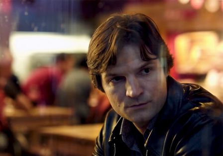 Ein Mann mit dunkelbraunem Haar sitzt in einem Restaurant, sein Gesicht ist ernst und konzentriert. Die Atmosph&auml;re ist dunkel und still.