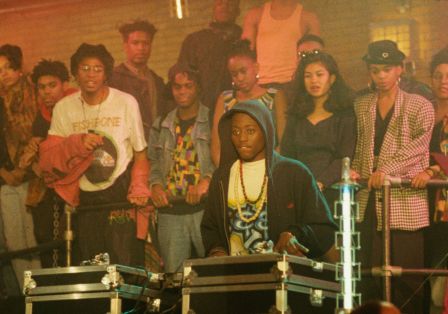 Ein junger Schwarzer Mann sitzt an DJ-Turntables, hinter ihm eine Gruppe von jungen Leuten, die ihm zuh&ouml;rt.