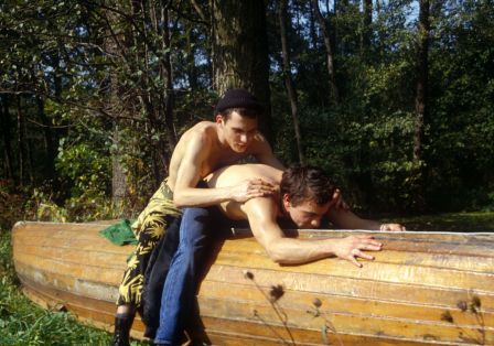 Zwei M&auml;nner mit freiem Oberk&ouml;rper sitzen auf einem umgedrehten Holzboot an Land, einer beugt sich &uuml;ber den anderen.