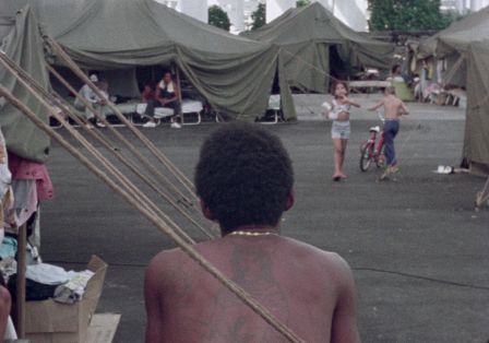 Ein schwarzer Mann mit Afro sitzt mit dem R&uuml;cken zur Kamera. Er schaut auf auf ein Zeltlager. 