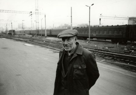 Ein alter Mann steht auf einem Bahnhofsvorplatz, mit einem Zug im Hintergrund.