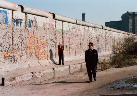 Mann im schwarzen Anzug und Sonnenbrille geht entlang der graffitibedeckten Berliner Mauer, während ein anderer in einem leuchtend Anzug im Hintergrund steht.