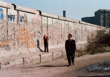 Mann im schwarzen Anzug und Sonnenbrille geht entlang der graffitibedeckten Berliner Mauer, w&auml;hrend ein anderer in einem leuchtend Anzug im Hintergrund steht.