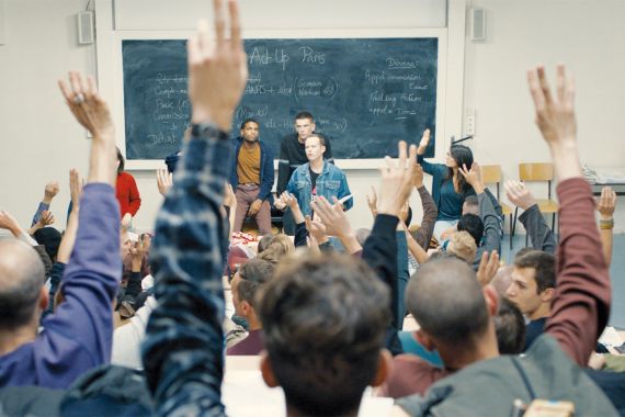 Viele H&auml;nde in einem Klassenzimmer, die hochgehen. An der Tafel stehen drei junge M&auml;nner.