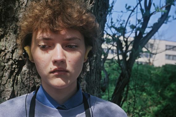 Eine Jugendliche in Nahaufnahme steht vor einem Baum, tr&auml;gt gelbe Kopfh&ouml;rer und blickt ernst zur Seite. A close-up of a teenage girl standing in front of a tree, wearing yellow headphones and gazing to the side.