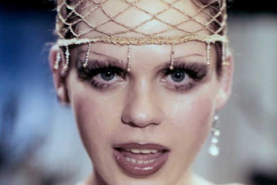 Eine stark geschminkte Frau blickt singend in die Kamera. A heavily made-up woman looks singing into the camera.