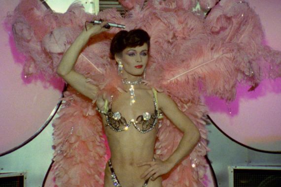 Eine Transperson in glitzerndem BH und mit Federn am Rücken macht eine Tanzbewegung. A transgender person in a glittery bra and with feathers on their back makes a dance move.