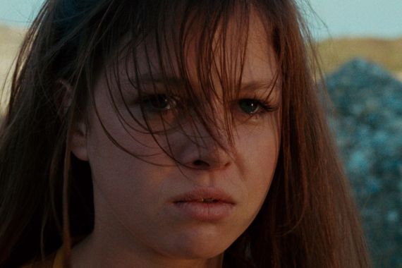 Nahaufnahme einer jungen Frau mit langem Haar im Wind, ernst blickend, mit verschwommener Felsenlandschaft im warmen, nat&uuml;rlichen Licht.