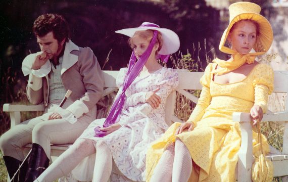 Drei Menschen in Sommerhüten, Kleidern und Jacke sitzen auf einer Bank und schauen mürrisch und lädiert vor sich.