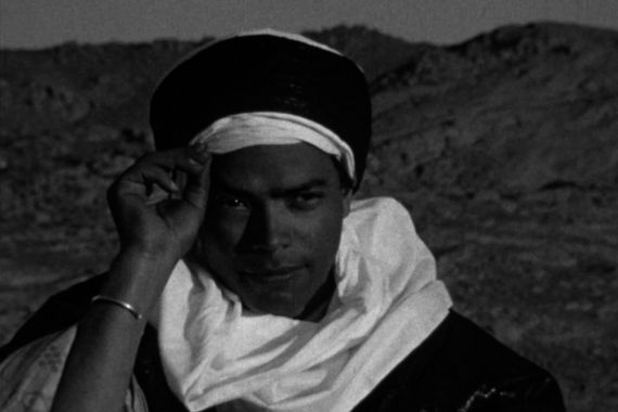 A man in a desert landscape is holding his turban and smiling.