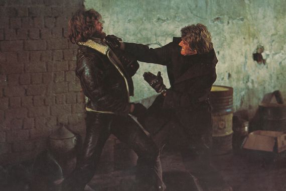 Zwei M&auml;nner in dunklen M&auml;nteln und Lederjacken mit halblangen Haaren k&auml;mpfen miteinander.