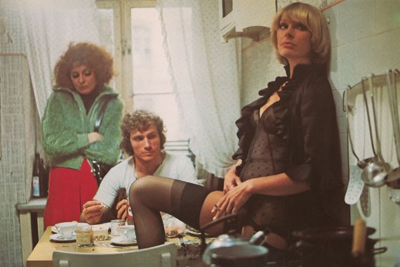 Ein Mann in einer K&uuml;che blickt auf das Bein einer Frau in Strapsen, w&auml;hrend die zweite Frau neben ihm auf den Boden schaut.