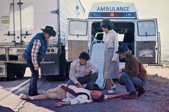  Vier Personen stehen um einen verletzten Mann auf dem Boden, hinter ihnen ist eine Ambulanz.