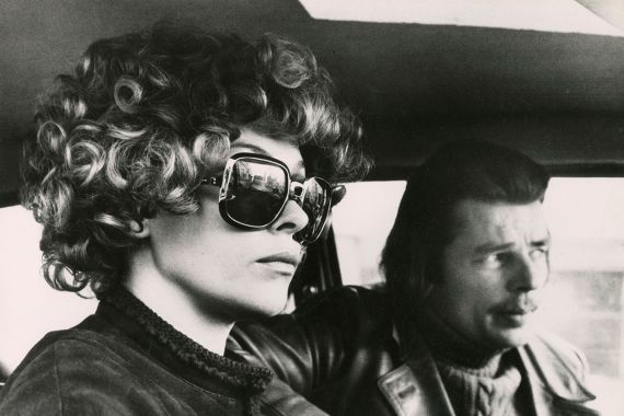 Eine Frau mit gro&szlig;er Sonnenbrille sitzt ernst neben einem Mann mit halblangem Haar und Schnurrbart im Auto.