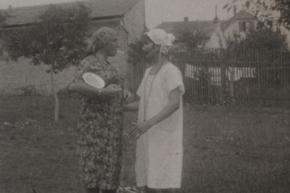 Zwei Männer in Kleidern und Hauben stehen in einem Garten und scheinen zu diskutieren.