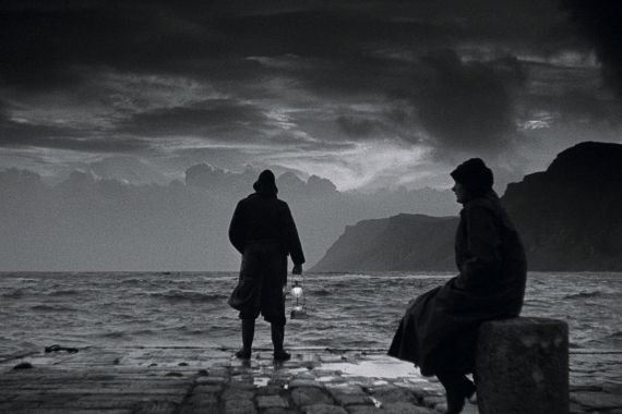 Eine Frau und ein Mann, beide in Regencapes, stehen am Meeresufer und blicken auf einen st&uuml;rmischen Himmel voller Wolken.
