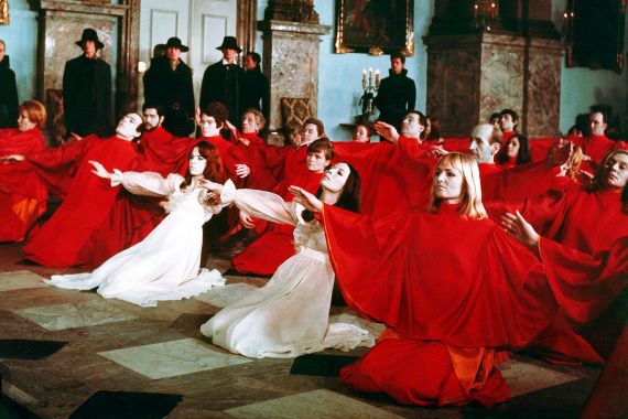 In einem schlossartigen Saal knien Frauen und M&auml;nner in roten und wei&szlig;en Gew&auml;ndern auf einem Marmorboden, w&auml;hrend M&auml;nner in Schwarz mit H&uuml;ten im Hintergrund stehen.