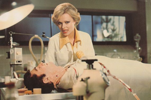 Eine Frau mit blonden Haaren und Vampirz&auml;hnen beugt sich &uuml;ber einen Mann in einem OP-Saal und faucht in die Kamera.