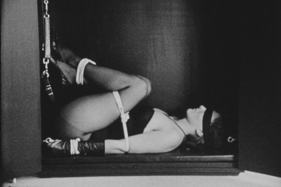 A blindfolded woman is lying in a box, with her arms and legs tied in a bondage style.