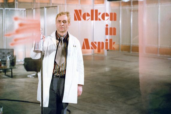 Ein Mann in einem Doktorkittel hält eine Farbsprühflasche und blickt in die Kamera.