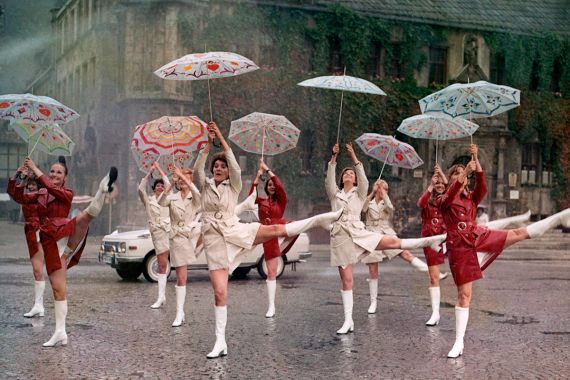 Eine Gruppe von Frauen in Regenmänteln und weißen Stiefeln tanzt mit Regenschirmen auf der Straße.