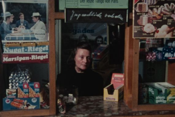 An elderly woman sits at a kiosk window, looking distressed.