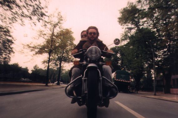 Ein Mann und eine Frau mit Sonnenbrillen fahren auf einem Motorrad frontal auf uns zu.