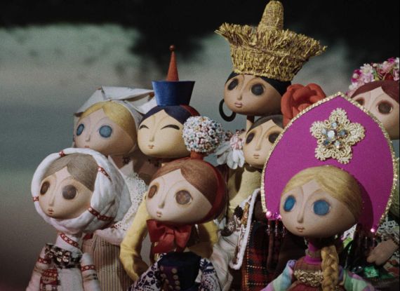 Szenenfoto aus einem Puppentrickfilm: Eine Gruppe von jungen Frauen in folkloristischer Kleidung blickt nach links auf ein Ereignis au&szlig;erhalb des Bildraums.