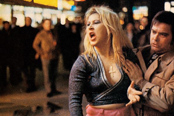 Eine Frau mit langen blonden Haaren schreit jemanden an und wird dabei festgehalten.