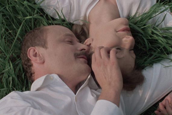 A man and a woman in white clothing lie side by side on a meadow, smiling at each other and touching.