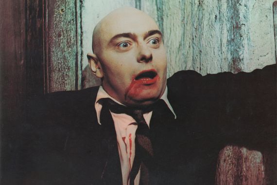  Ein Mann ohne Haare mit weit aufgerissenen Augen hat einen blutverschmierten Mund und Blut auf seinem weißen Hemd.