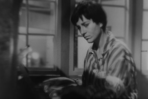 A dark-haired woman in prisoner’s clothing plays the piano and looks pensively to the side.