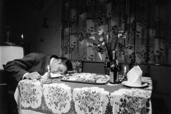 A man is sleeping with his head on the kitchen table, next to him are flowers, champagne, candles, and a plate of food.