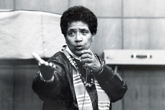 Eine schwarze Person mit Afro, Jacke und Kette, spricht in ein Mikrofon in ihrer Hand. Mit der anderen Hand zeigt sie nach vorne.