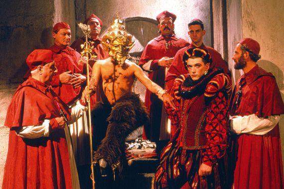 7 M&auml;nner in roten Gew&auml;ndern um einen nackten Mann versammelt, der eine goldene Maske tr&auml;gt.
