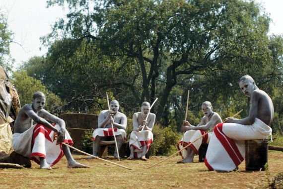 Gruppe von schwarzen M&auml;nnern in traditioneller Kleidung sitzt im Freien. Sie sind mit wei&szlig;er Farbe bemalt.