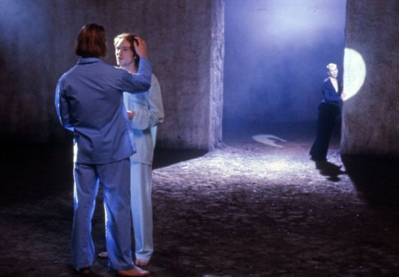 Zwei Personen in blauen Anz&uuml;gen stehen auf einem blau beleuchteten Platz. Eine andere Person ist im Hintergrund.