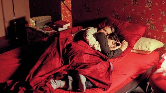 Zwei Personen liegen umschlungen in einem Bett. W&auml;nde, Bett und Decke sind rot.