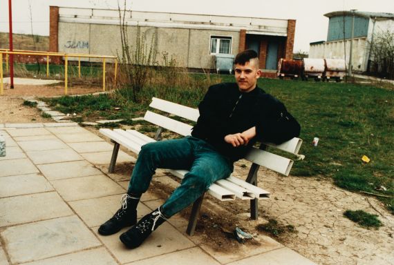Ein junger Mann sitzt auf einer Bank. Seine Haare sind an den Seiten rasiert, er tr&auml;gt eine Bomberjacke, Jeans und Springerstiefel.