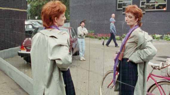 Eine Frauen mit roten Haaren und 80er Jahre Style betrachtet sich in einem Spiegel auf der Stra&szlig;e. 