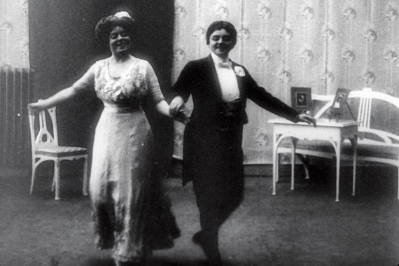 Schwarz-wei&szlig;-Filmstill einer historischen Szene: Zwei Frauen (eine im Kleid, eine im Anzug) tanzen in einem Zimmer.