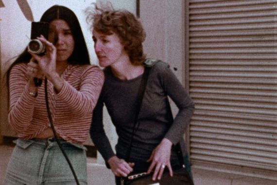 Two women, one with long black hair and the other with red curls, are looking through a camera and have a recording device with them.