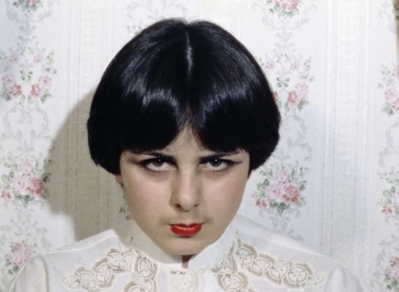 M&auml;dchen mit Bob und rotem Lippenstift schaut intensiv in die Kamera. Hinter ihr eine altmodische Tapete mit Blumenmuster