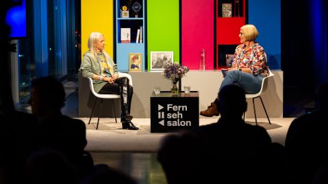Zwei Frauen unterhalten sich in einem Studio, das in den grellen Farben eines Fernseh-Testbilds gestaltet ist.