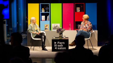 Zwei Frauen unterhalten sich in einem Studio, das in den grellen Farben eines Fernseh-Testbilds gestaltet ist.