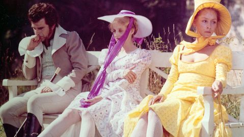 Drei Menschen in Sommerhüten, Kleidern und Jacke sitzen auf einer Bank und schauen mürrisch und lädiert vor sich.