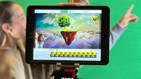 Kinder hinter einem Tablet auf einem Stativ, vor einem Greenscreen. Auf dem Tablet sind die Kinder in einer Landschaft zu sehen.