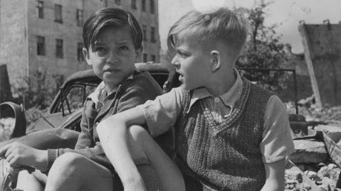 Zwei Jungen in kurzer Kleidung sitzen auf Tr&uuml;mmern in kriegszerst&ouml;rtem Berlin, mit Autowrack und zerst&ouml;rten H&auml;usern im Hintergrund.